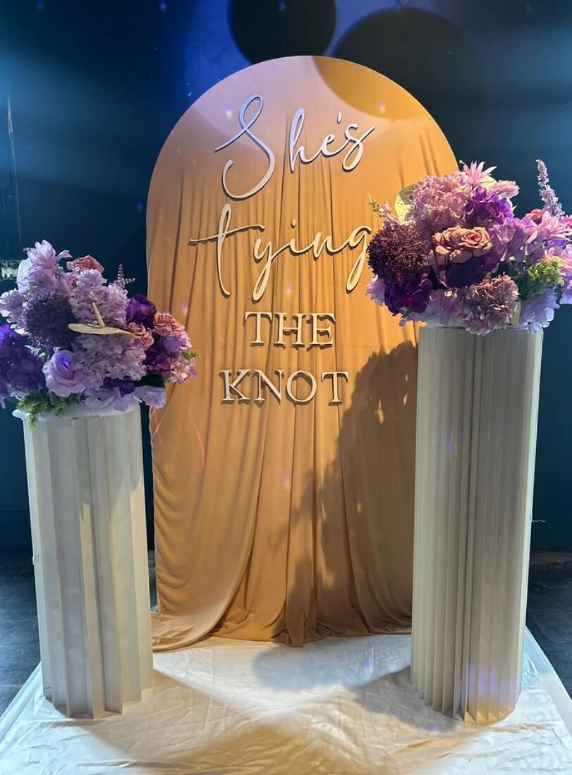 Gold draped arch backdrop with acrylic lettering and purple floral plinths